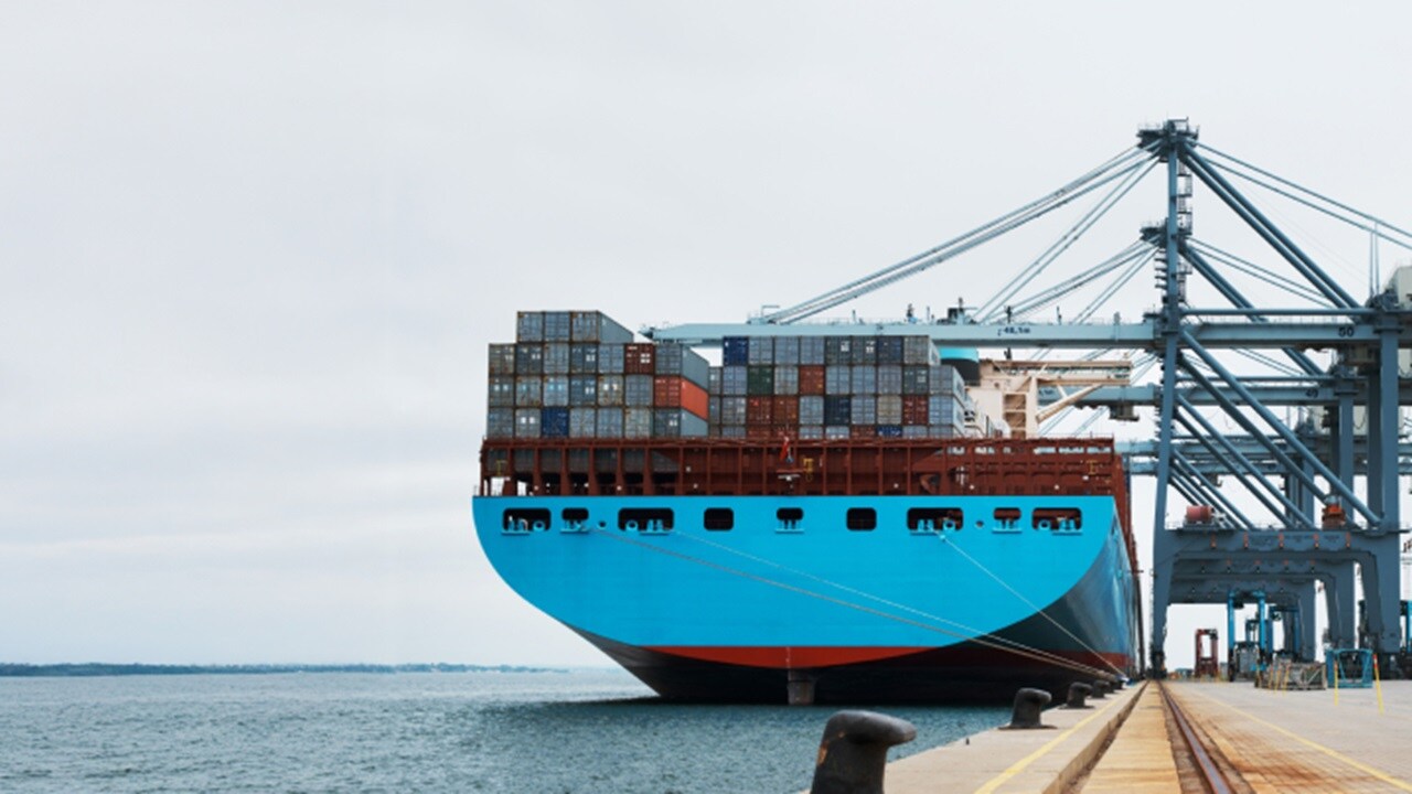 A large container ship is docked at a port, with cranes and cargo containers visible in the background.