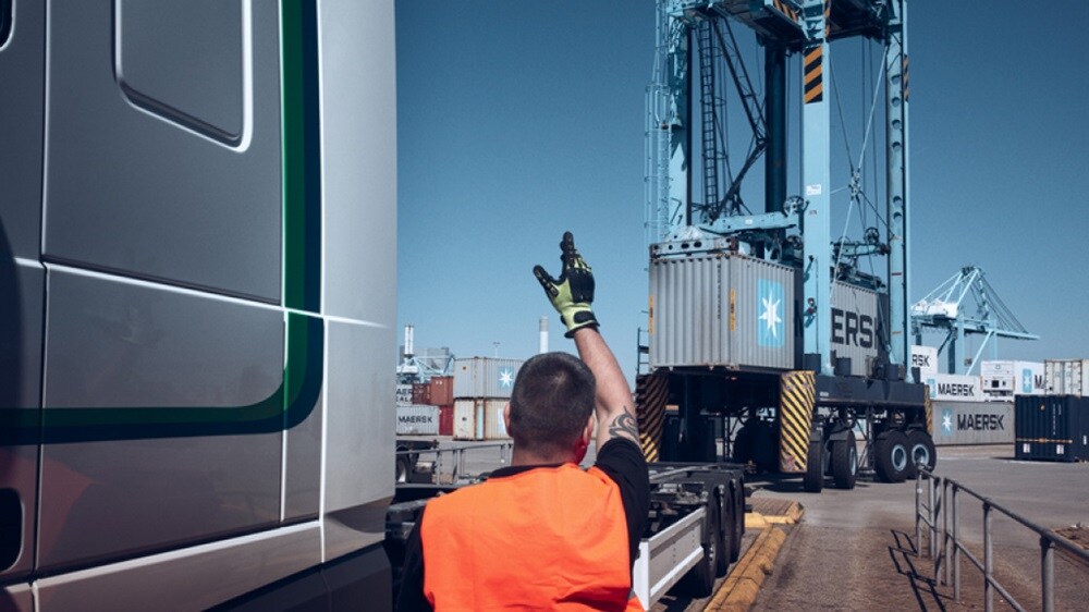 A logistics worker in an orange safety vest signaling to a truck driver near a large Maersk shipping container at a container terminal.