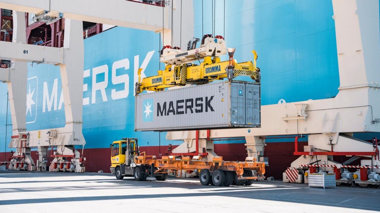 A Maersk shipping container being loaded onto a truck by a large crane at a container terminal, with a Maersk ship in the background.