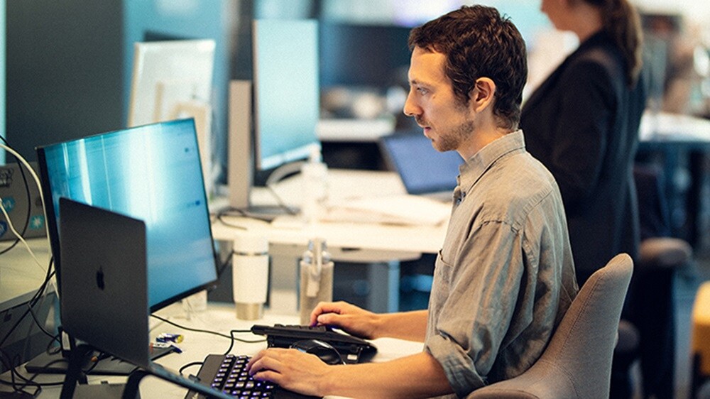 An engineer at his workstation with two computers and typing on keyword with a woman in the background.