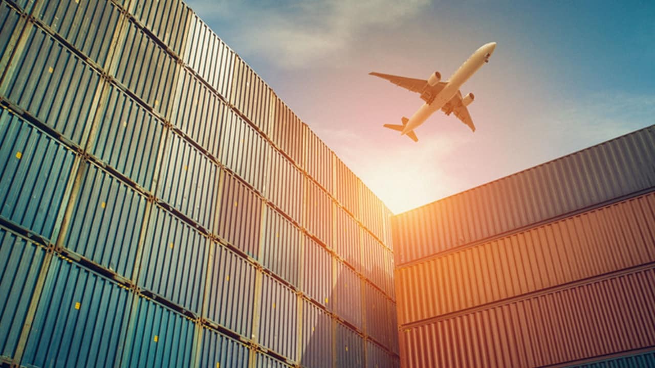Walls of multi-coloured, stacked shipping containers are pictured against a blue sky with a plane flying overhead. 