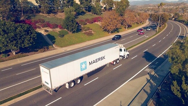 A semi truck drives along a highway, with green trees lining the background under a clear blue sky.