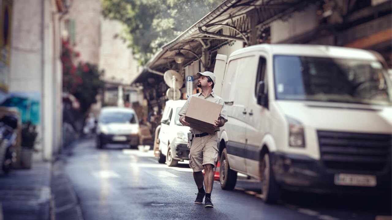 A delivery man wearing beige shorts and a matching shirt is holding a package and walking down a street looking at house numbers. His white delivery van is visible in the background.