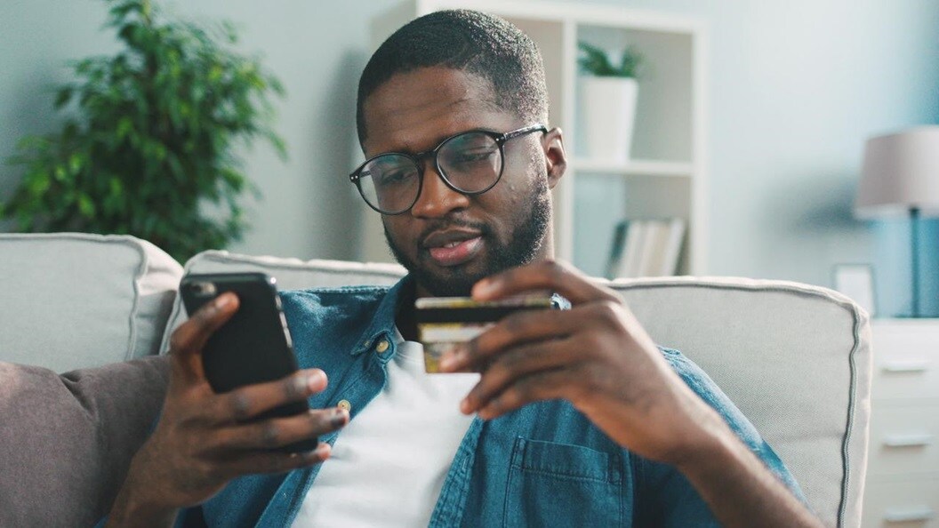 A black man sitting on a cream couch. He is holding his phone in one hand and looking at the screen. In his other hand he is holding up a credit card to read the numbers.  
