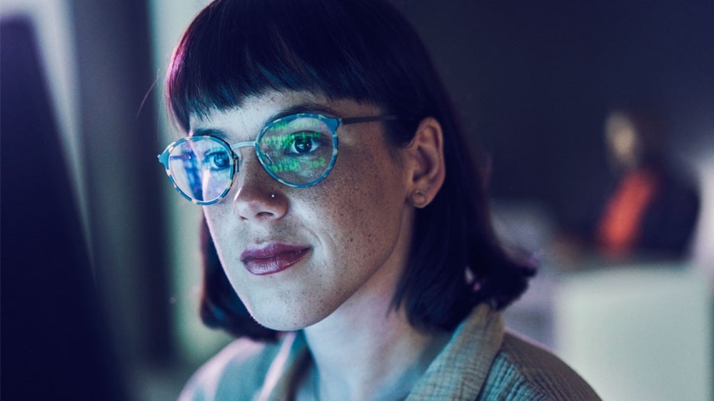 A woman wearing glasses looking into a screen on which artificial intelligence code is shown.