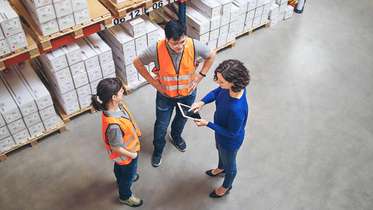 Two workers in safety vests and helmets handling cardboard boxes near a delivery truck in a warehouse.