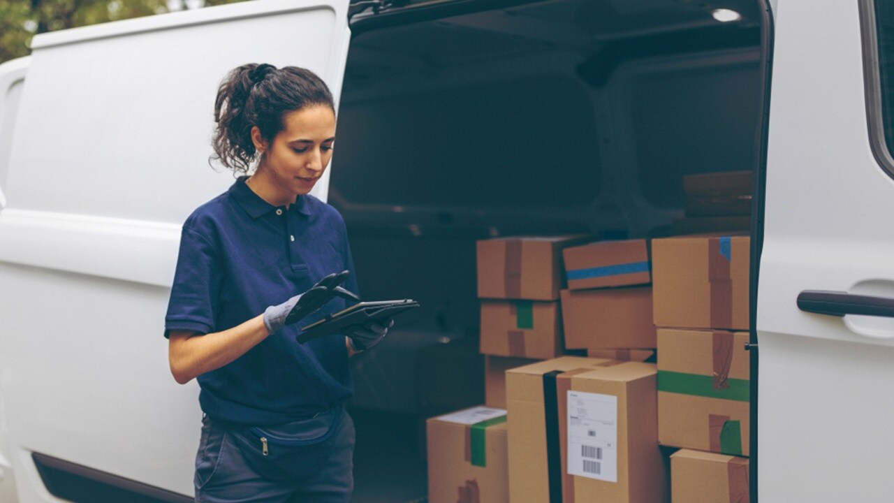 A delivery person with an obscured face stands next to an open van, holding a handheld device, with multiple cardboard boxes in the background.