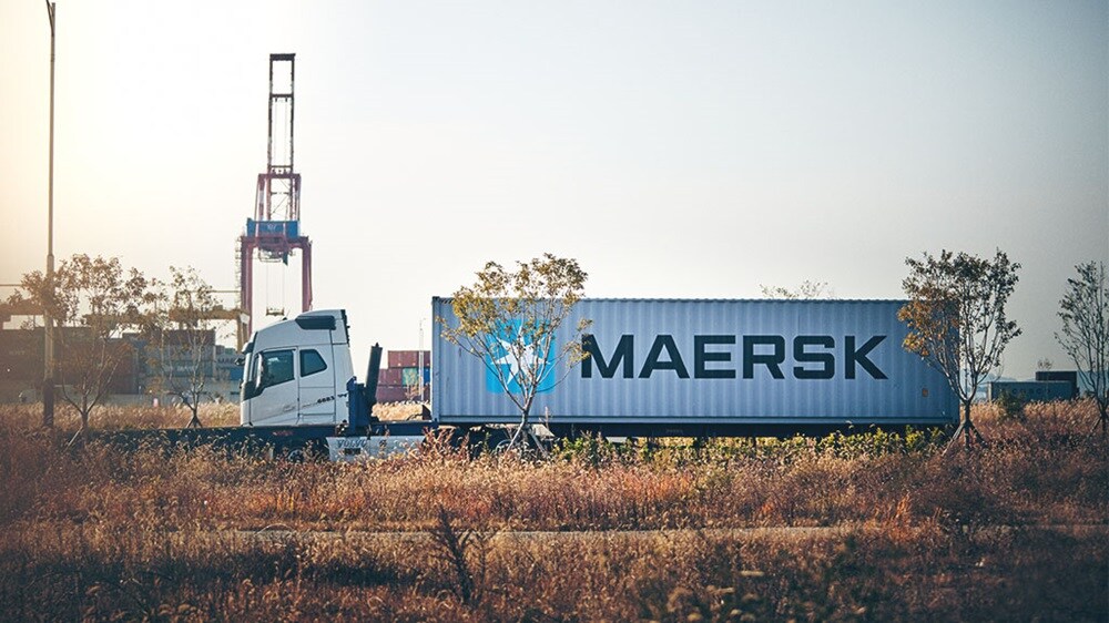 A semi-truck with a white trailer bearing the ‘MAERSK’ logo is driving under an overpass on a clear day.