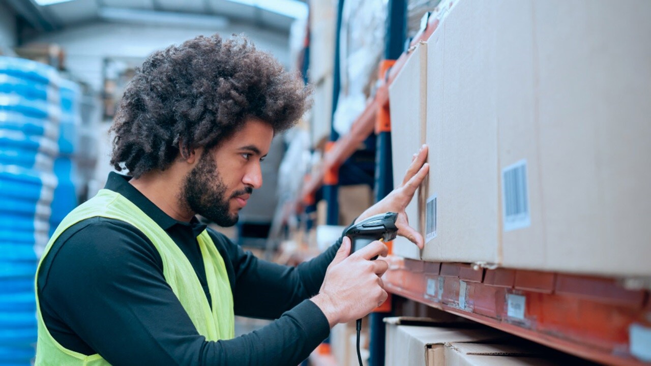 A person in a high-visibility vest using a handheld barcode scanner to scan items on a warehouse shelf.