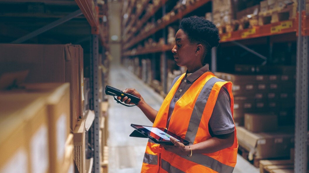 A person holding a clipboard and a handheld device stands in a warehouse aisle, checking inventory.