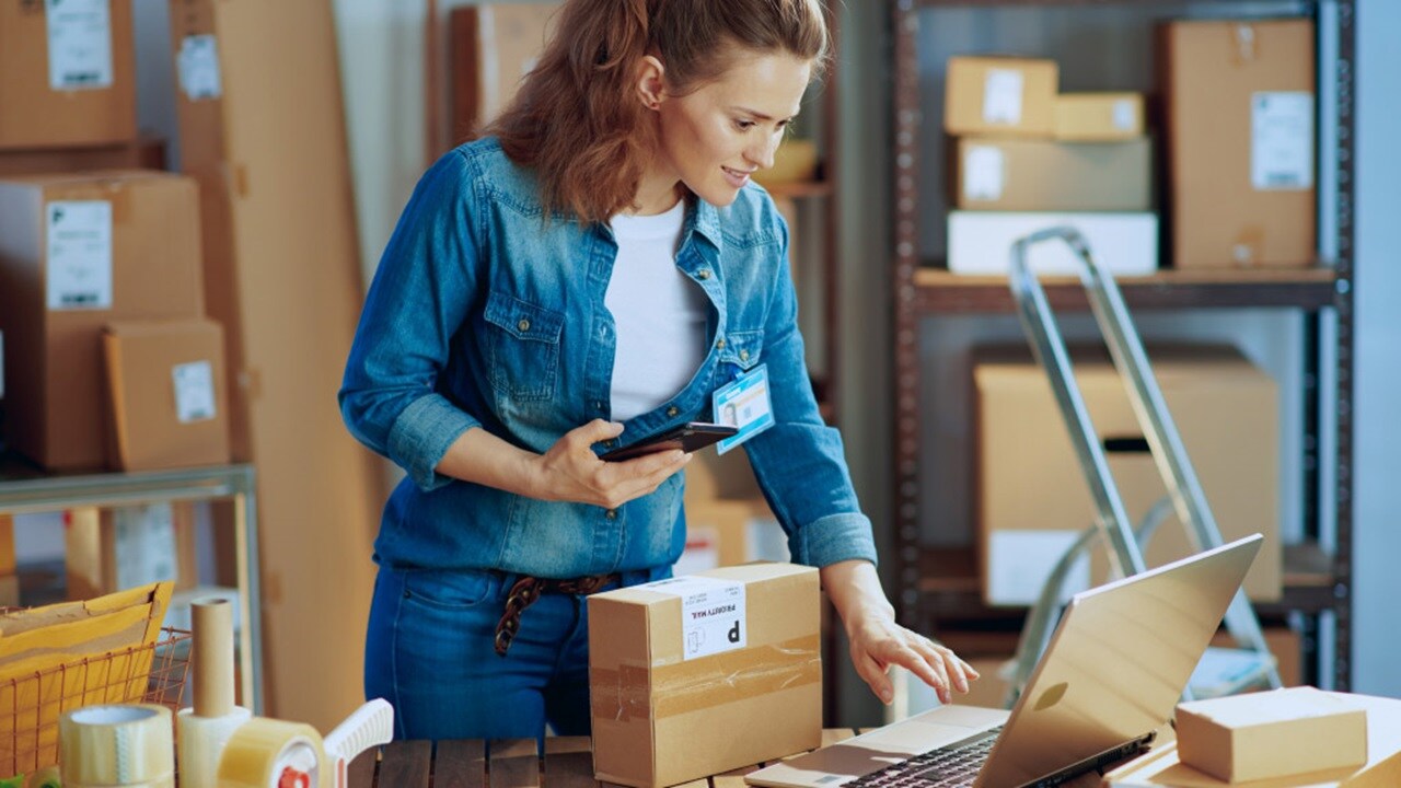 A person in a blue shirt and jeans stands in a warehouse, using a laptop placed on a table with various packages around. The person is holding a handheld device, possibly for inventory management or scanning items.