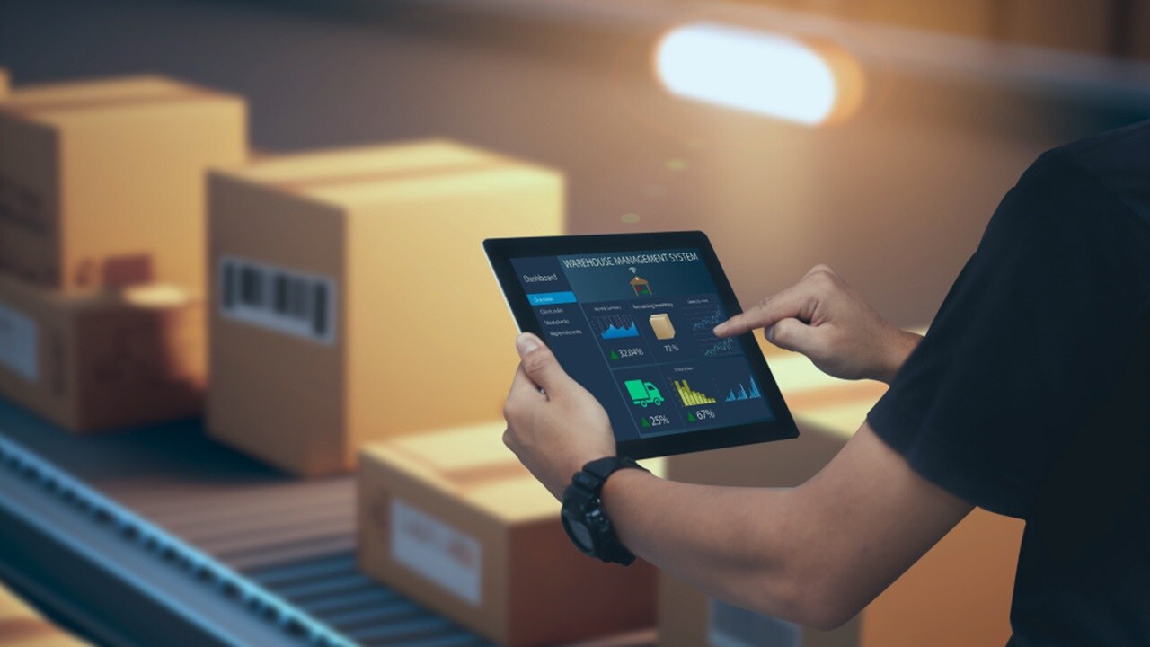 A person holding a digital tablet with logistics graphics on the screen, standing in a warehouse with packages in the background.