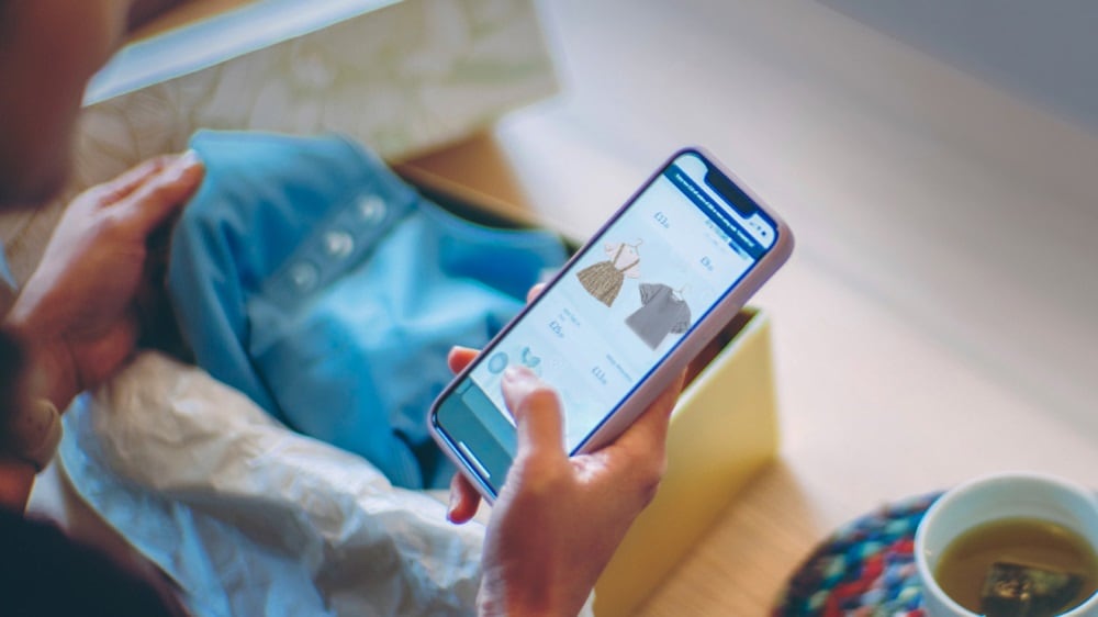 A person holding a smartphone with an online shopping application open, displaying a piece of clothing for sale. The individual is seated, with a cup and saucer visible on the table beside them.