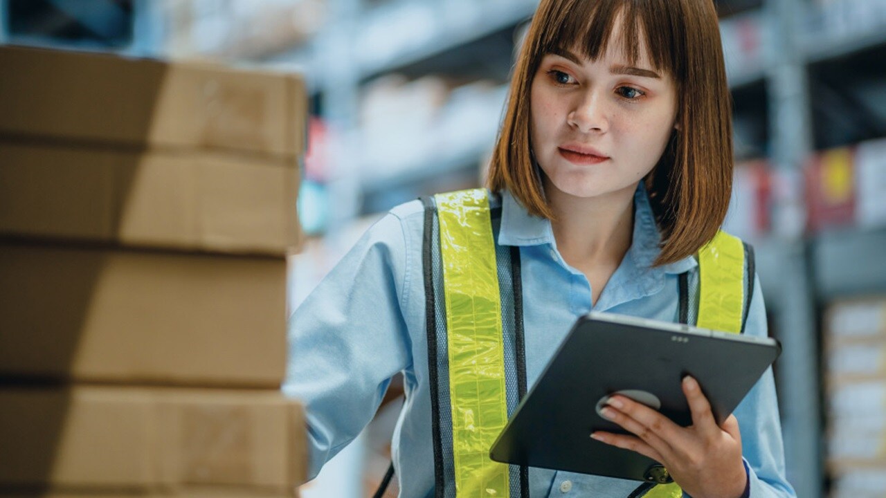 An individual in a high-visibility vest holding a clipboard in a warehouse with boxes in the background.