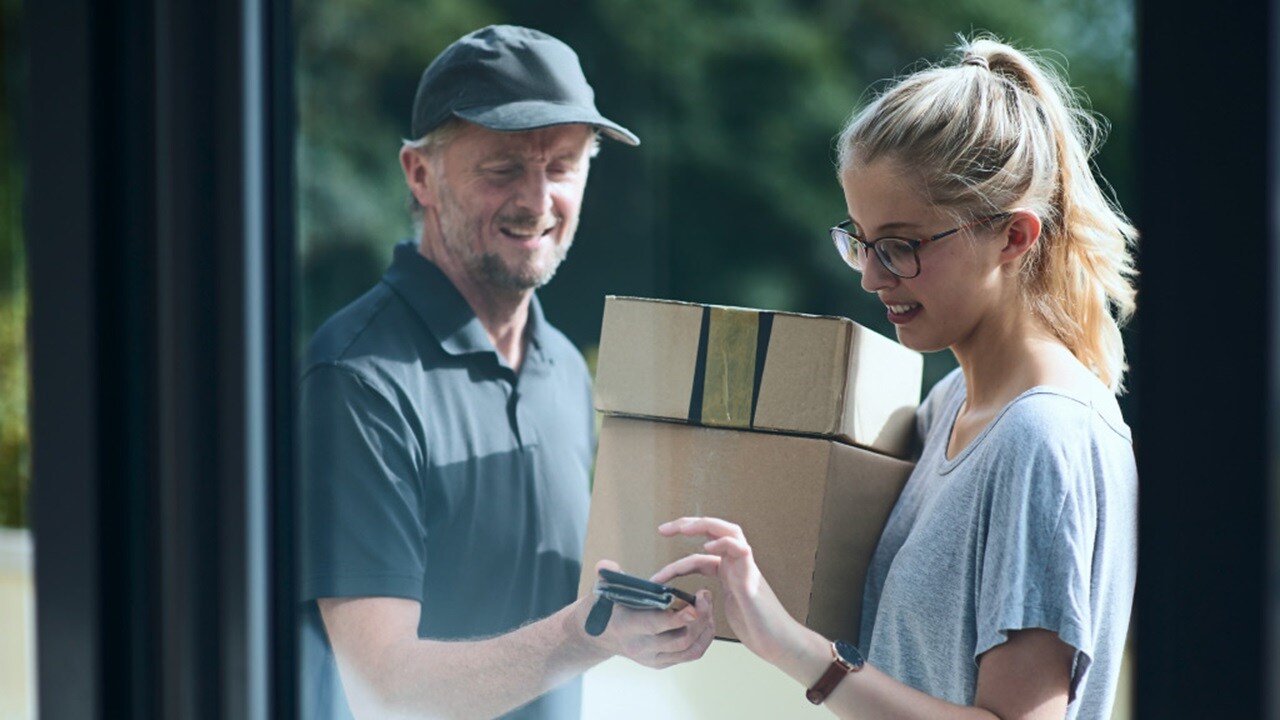 A person in a black cap handing over a stack of boxes to another person holding a mobile device, with their faces obscured by brown rectangles.