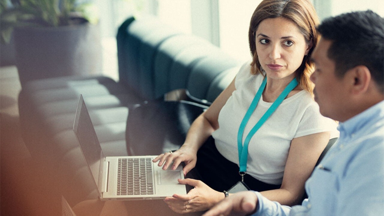 Two people – a woman and a man- dressed in office clothes are sitting next to each other on a couch. The man is looking at the screen of a computer on the woman’s lap. She is looking at his face while he talks. 