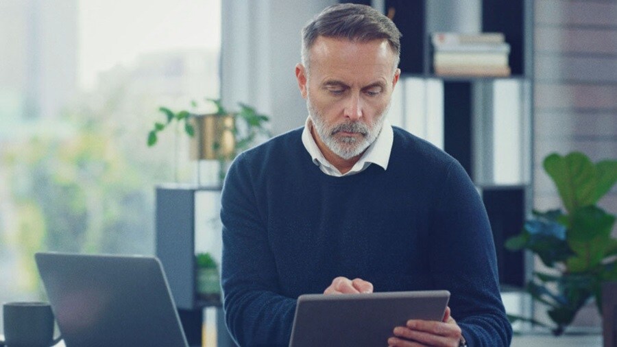Focused man using a tablet in a modern office with a laptop and plants in the background.