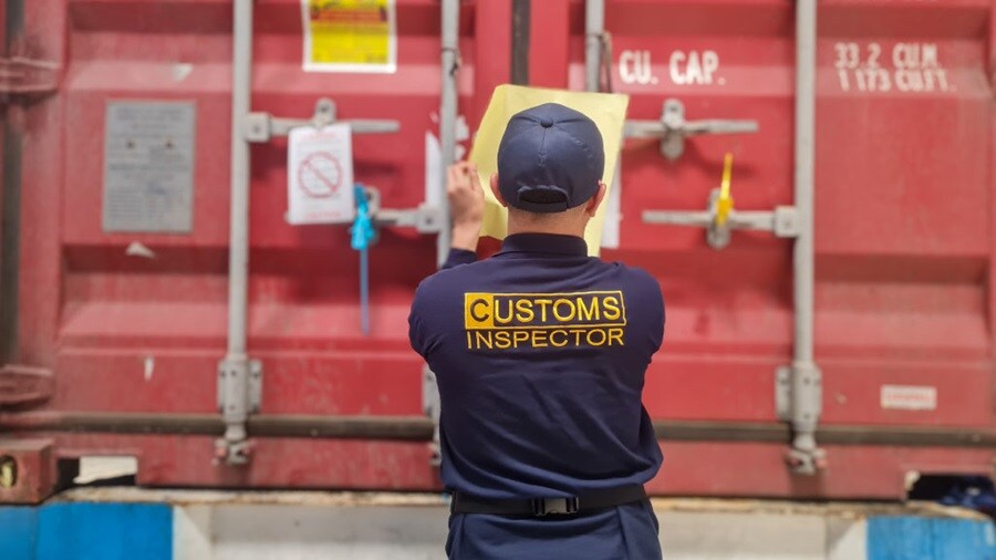 A customs inspector in uniform affixes a document to a sealed red shipping container.