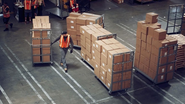 A warehouse worker in a safety vest pushing a cart loaded with boxes alongside organized pallets in a logistics facility