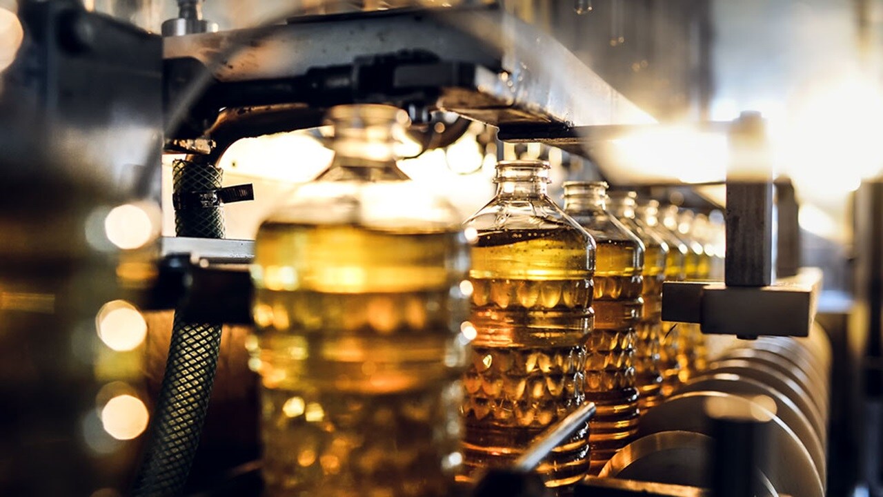 A row of ornate glass bottles on a factory line are being refilled with a thick yellow liquid. 