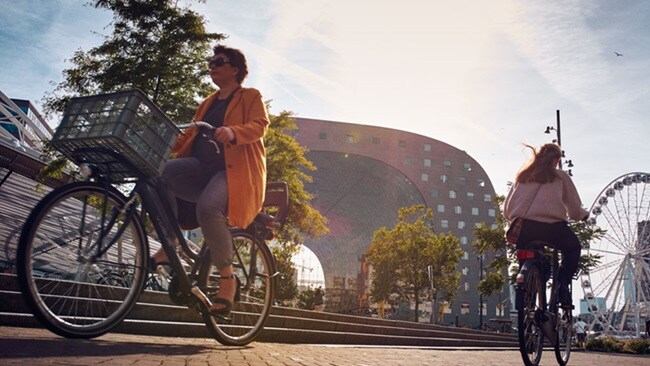 Two women on bicycles cycle away from each other. In the background is a Ferris wheel and a building in the shape of an arch.
