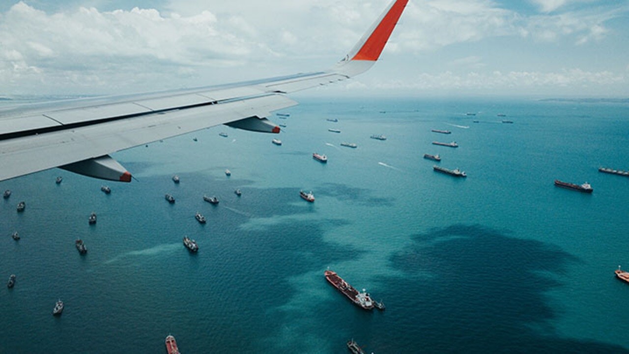 Wing of airplane flying over container ships on the ocean