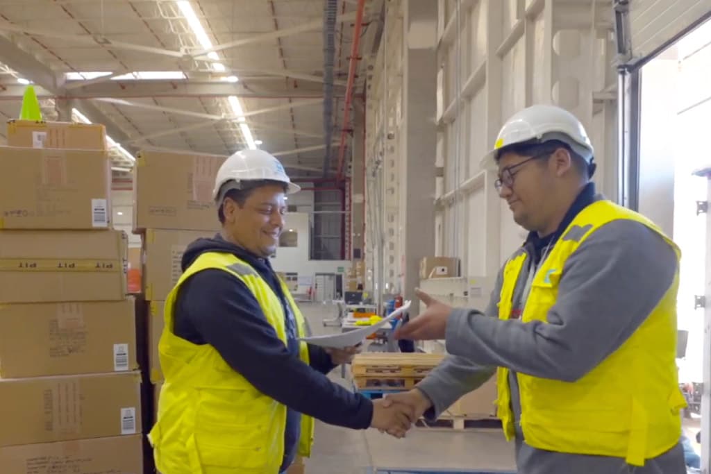 Two warehouse workers in safety vests and helmets shake hands while exchanging documents.