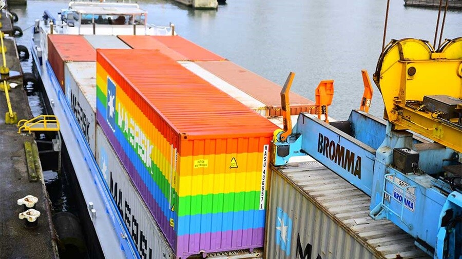 Maersk shipping containers, including a rainbow-colored one, on a barge at a port.