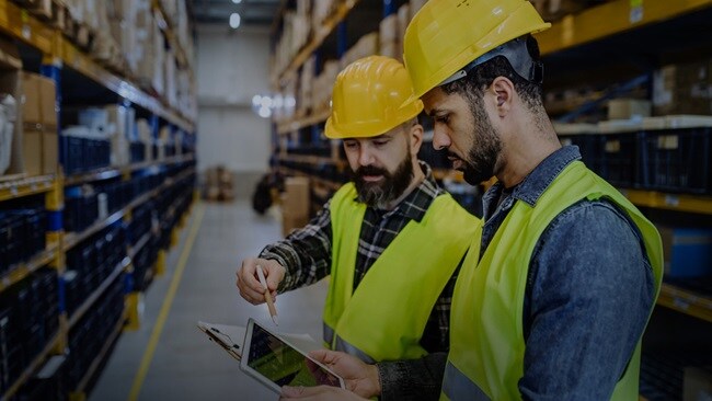 Two persons wearing yellow jackets and yellow hats in a warehouse