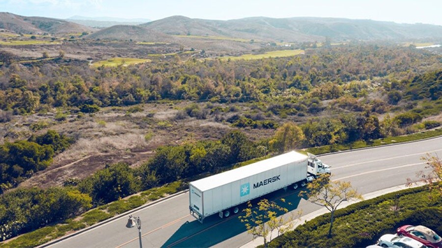 A Maersk truck drives along a scenic, winding road surrounded by hills and greenery.