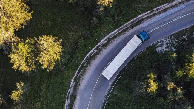 Aerial view of a truck traveling along a winding road surrounded by greenery and open fields.