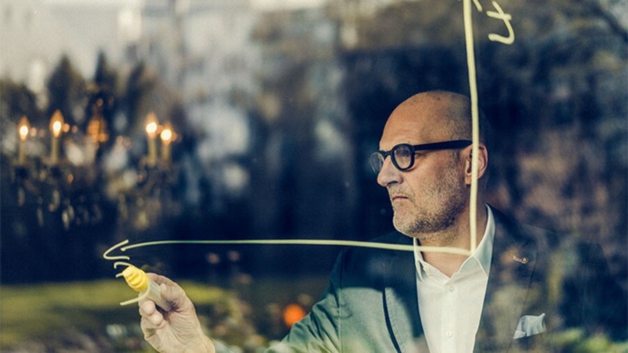 Man drawing a graph on glass with a yellow marker, seen through a reflective window.