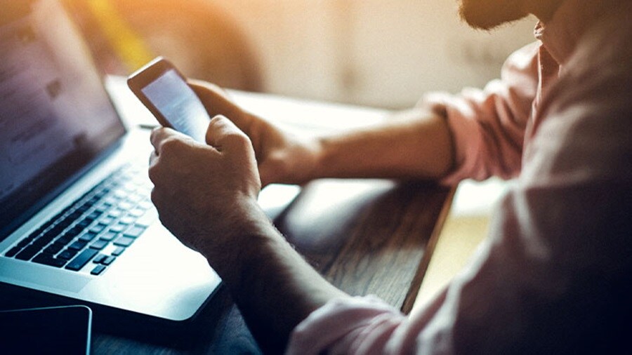 Man using a smartphone while working on a laptop at a sunlit desk.