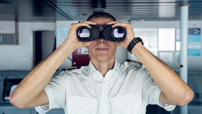 A ship officer in uniform uses binoculars to scan the horizon from the bridge.