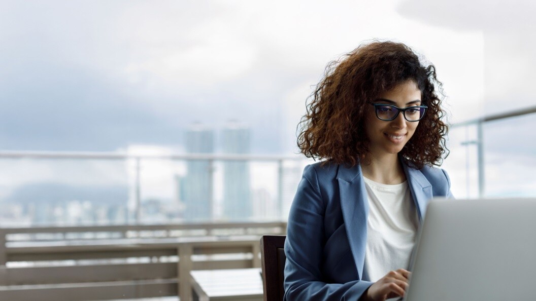 A professional woman wearing glasses and a suit is typing on her laptop, indicating a work-related task.