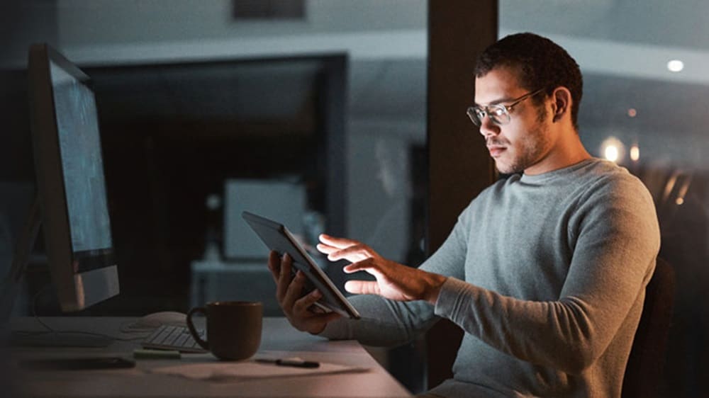 A man with glasses seated at their workstation and using a tablet to explore digital solutions.