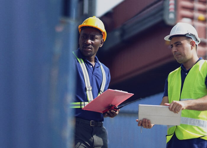 A couple of male workers in protective gear looking at a cargo container at a terminal.