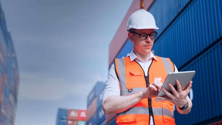 A man in an orange safety vest and hard hat stands with a tablet, engaged in tasks at a construction site.