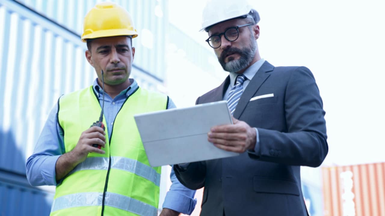 Two hard-hatted men stand next to a container, likely discussing logistics or safety measures in a construction environment.