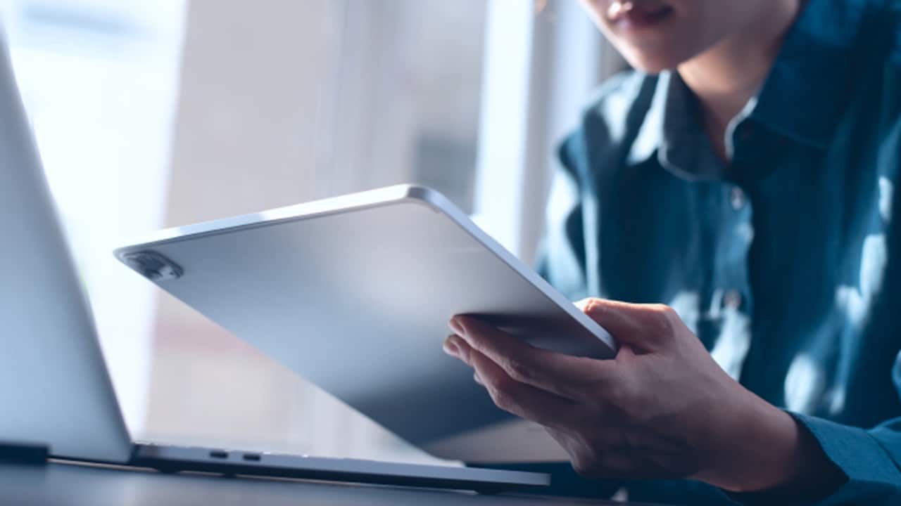 Person using a tablet while working on a laptop at a bright desk near a window.