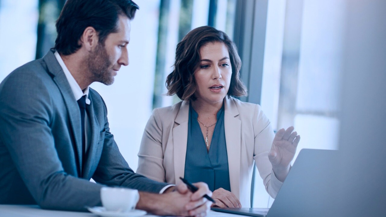Two professionals in business attire discuss something on a laptop screen in a bright office setting.