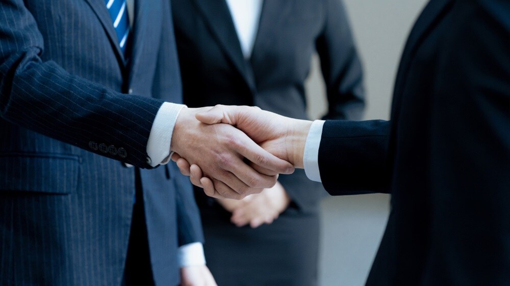 Two business professionals in suits shake hands, symbolizing a formal agreement, with a third individual in a suit in the background.
