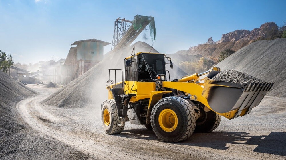 A yellow front-end loader is actively moving gravel at a construction site, surrounded by piles of material under a clear blue sky.