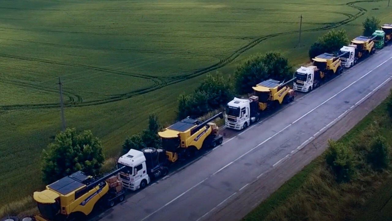 Trucks transporting yellow harvesters on a rural road near farmland.
