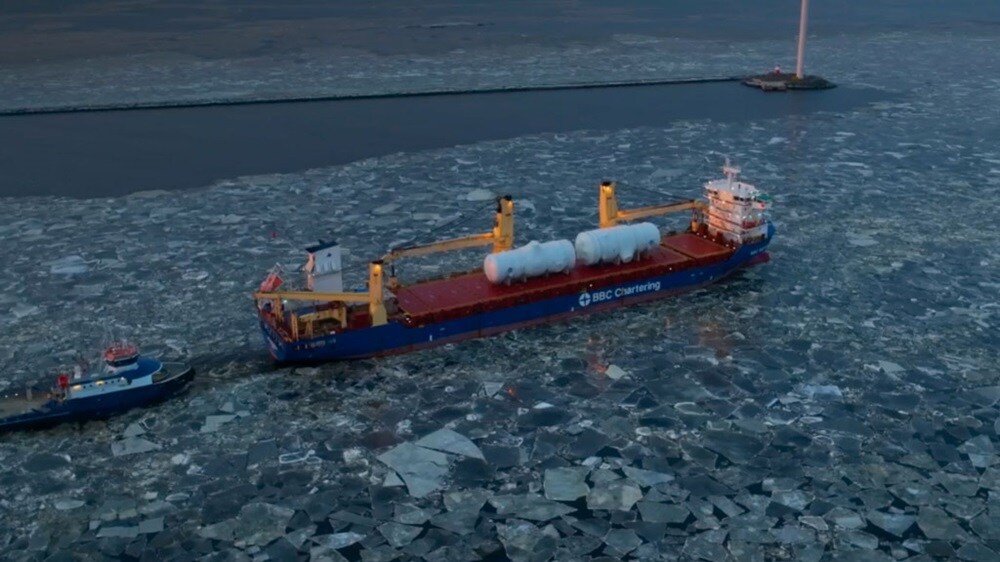 A cargo ship from BBC Chartering carrying large industrial equipment navigates through an icy waterway, escorted by a tugboat.