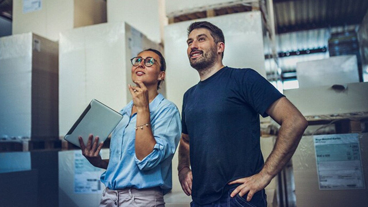 Two people, one holding a tablet, stand in a warehouse with large boxes.