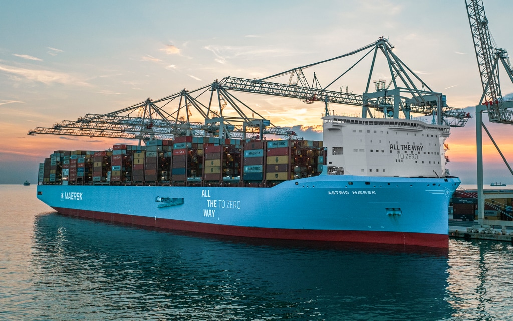 A Maersk container ship filled with stacked cargo containers is docked on the ocean.