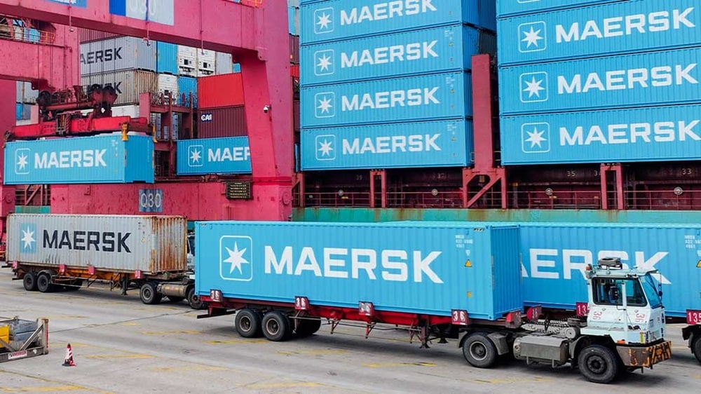 Maersk container being loaded onto a truck at a port terminal using a gantry crane, surrounded by stacked shipping containers