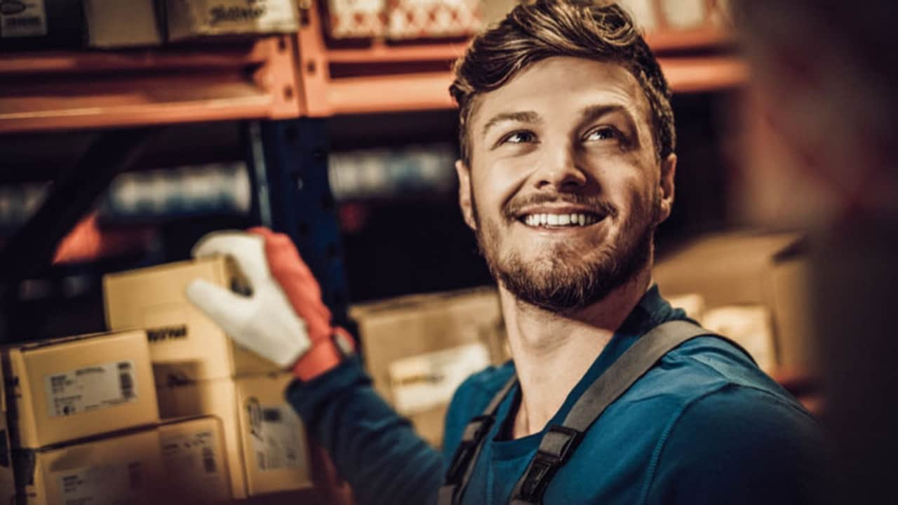 Close-up of a smiling man looking sideways and holding a small cargo box in a warehouse.