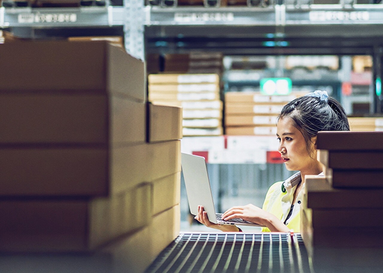 A woman looking at her laptop in a warehouse containing small boxes. 